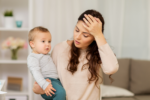 Parental burnout: Mother holding baby while resting her hand against her forehead in show of discomfort