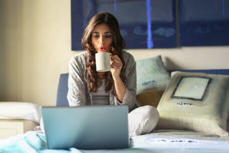 Woman blowing into coffee cup looking at open laptop: spend less time side hustle