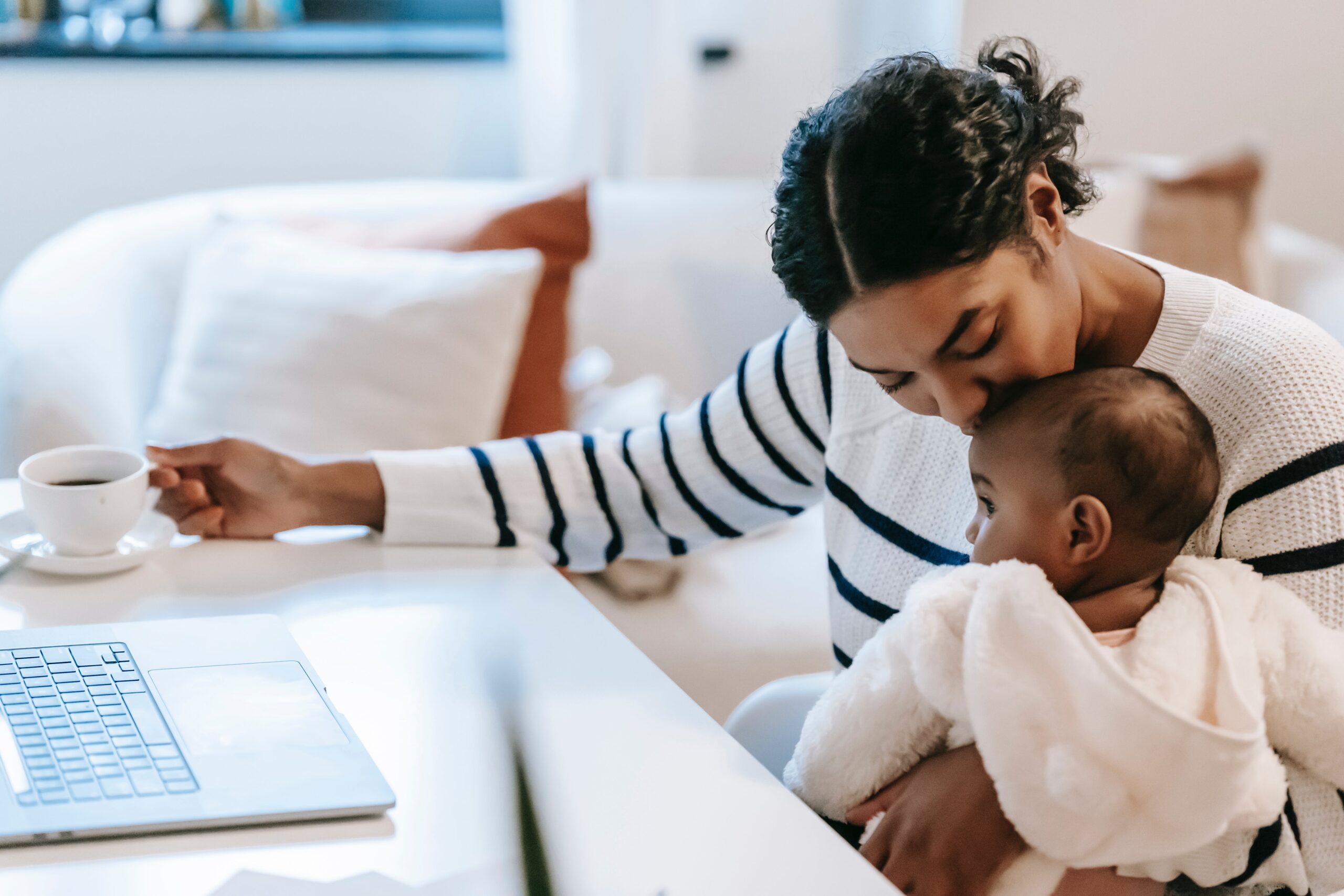 Ethnic woman kissing-baby near netbook and coffee cup: Navigating goals with new baby